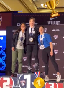 Professor Katie Rose Hejtmanek (center) stands on the podium with her first-place medal at the USA Weightlifting Masters National Championships.