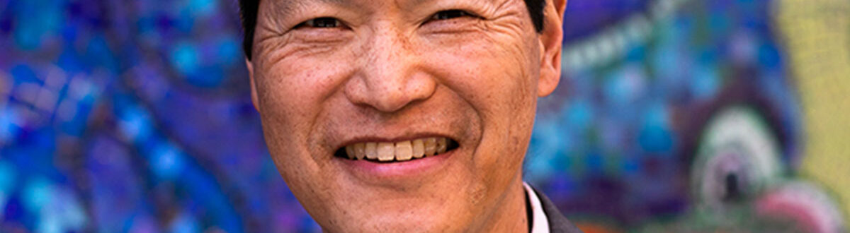 A close-up of Russell Jeung smiling wearing a light-colored button-down shirt and a gray suit jacket. The background is a colorful, textured mosaic with shades of blue, purple, and green.